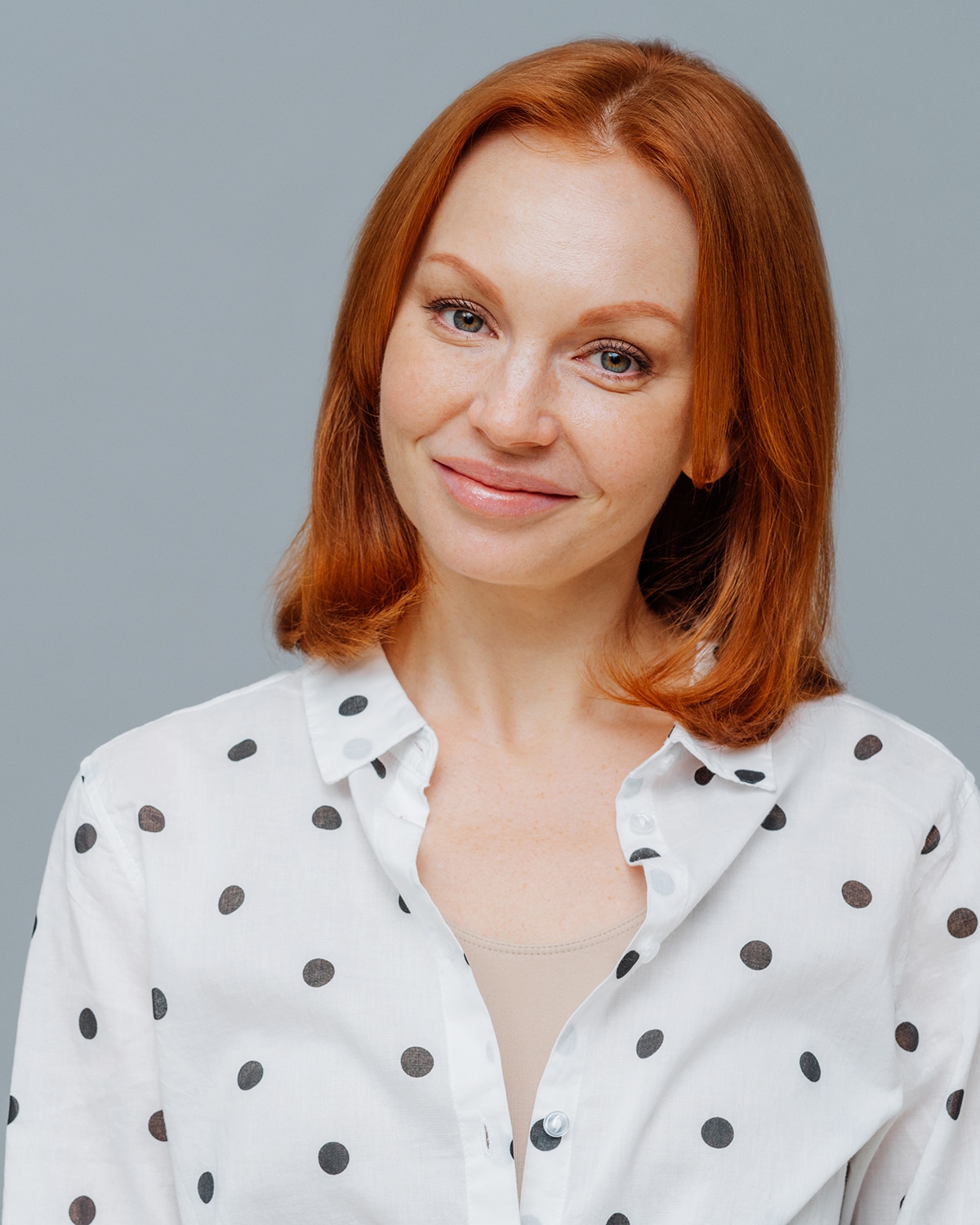 Pleased businesswoman with short red hair, makeup, looks positively at camera, has good mood after successful business meeting, dressed in stylish shirt, isolated over grey studio background.